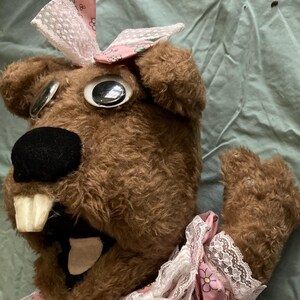 May include: A brown furry puppet with large eyes and a pink dress with lace trim and floral print. The puppet has a large mouth with white teeth and a pink tongue.