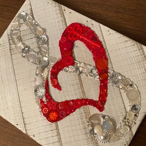 May include: A heart-shaped artwork made from red buttons and a silver infinity symbol made from buttons and beads. The artwork is mounted on a white-washed wooden plank background.