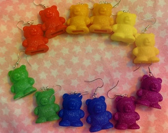 May include: A collection of colorful gummy bear earrings in a rainbow of colors: red, orange, yellow, green, blue, and purple. Each earring features a small, plastic gummy bear charm with a silver-tone hook.