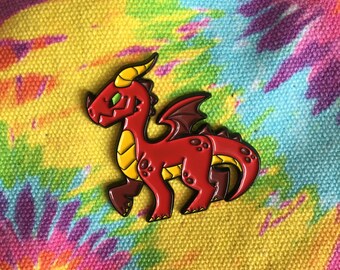 May include: A red dragon enamel pin with yellow accents and a black outline. The dragon has green eyes, yellow horns, and brown feet. The pin is set against a colorful tie-dye background.