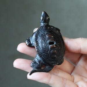 Puede incluir: Pequeña figura de tortuga marrón oscuro con patrones detallados en el caparazón y un agujero en el centro. La tortuga se sostiene en una mano, mostrando su tamaño y diseño intrincado. Estética clásica y antigua.