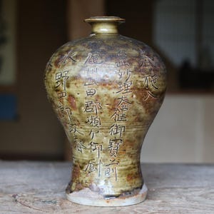 May include: A handcrafted, antique-style ceramic vase with a bulbous body and a narrow neck. The vase features a mottled, earthy color palette of brown, tan, and olive green. Intricate, stylized calligraphy decorates the surface.