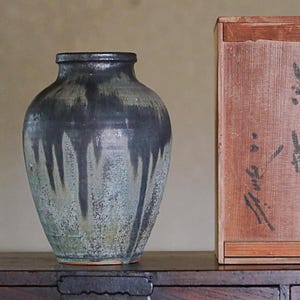 Puede incluir: Un jarrón de cerámica con un esmalte oscuro y moteado que presenta patrones de goteo verticales en tonos negro, gris y verde apagado. Junto al jarrón hay una caja de almacenamiento rectangular de madera con caracteres de caligrafía japonesa.