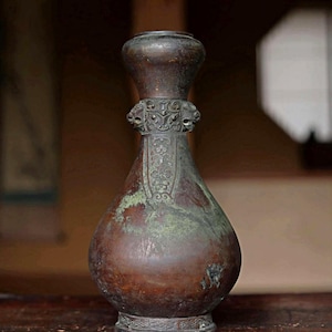 以下が含まれることがあります： 丸みを帯びた胴体と細い首を持つ茶色のアンティーク花瓶。 花瓶は首の周りに装飾的な彫刻が施され、風化した外観をしています。 花瓶の高さは約25cmです。