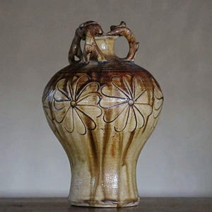 May include: A ceramic vase with a tan base and brown glaze drips. The body features carved floral patterns with multiple petals. Two sculpted handles shaped like small animals are attached to the neck of the vessel, which has a wide, rounded body.