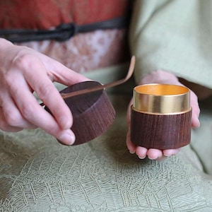 以下が含まれることがあります： 金色の内側と蓋が付いた木製の茶筒が開いています。茶筒は濃い茶色で、金色のバンドが付いています。木製のスプーンが蓋の上に置かれています。背景はぼやけています。