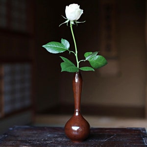 以下が含まれることがあります： 濃い茶色の陶器の花瓶に、白いバラが1輪と緑の葉が活けられています。花瓶は暗い木製のテーブルの上に置かれています。