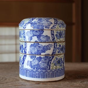Puede incluir: Un recipiente cilíndrico de cerámica de tres niveles con base blanca, decorado con patrones florales azules y diseños geométricos complejos. El recipiente apilable consta de tres secciones separadas con una tapa a juego, todas adornadas con motivos detallados de crisantemos azules.