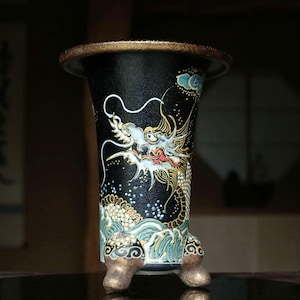 Könnte beinhalten: Eine schwarze zylindrische Vase mit breitem Rand und drei strukturierten goldfarbenen Füßen. Die Außenseite zeigt eine detaillierte Drachenillustration mit weißen, goldenen und roten Akzenten über stilisierten blau-weißen Wellen.