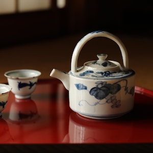 以下が含まれることがあります： 青い花柄の白い磁器の急須と、赤い漆塗りの盆の上にある2つの茶碗のセット