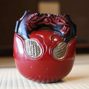 Puede incluir: Objeto decorativo de madera tallada en rojo y negro. El objeto tiene una base redondeada con una superficie texturizada y dos adornos dorados. La parte superior presenta un diseño negro tallado con dos figuras de dragón.