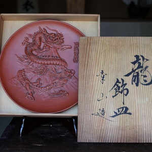 May include: A red ceramic plate with a raised design of a dragon on a mountain. The plate is in a wooden box with a Japanese inscription on the lid.