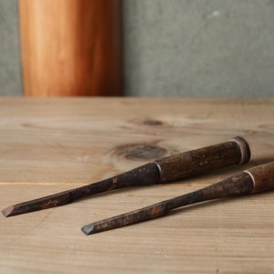 May include: Two wooden chisels with dark brown handles and metal blades lie on a light brown wooden surface.