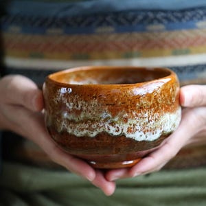May include: A handmade ceramic tea bowl with a brown and white glaze. The bowl is held in both hands, with a blurred background of a patterned textile and green fabric.