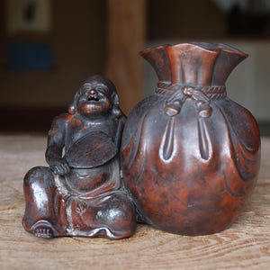 May include: A wooden sculpture of a seated, smiling figure holding a fan next to a round, bag-shaped container. The sculpture is a dark reddish-brown color and sits on a wooden surface. The figure and container appear to be part of a decorative set.