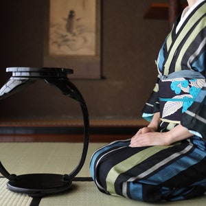 Puede incluir: Una mesa redonda de madera negra con un diseño único. La mesa está sobre una estera de paja y una persona vestida con un kimono a rayas azul y verde está sentada frente a ella.