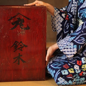 May include: A person wearing a blue and white patterned kimono sits on a straw mat with their hand resting on a wooden box. The box is dark red and has black Japanese characters on the front that read "珍藏木".