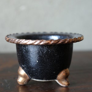 Puede incluir: Una pequeña maceta de cerámica negra con un borde dorado y tres patas doradas. La maceta es perfecta para plantas pequeñas o suculentas.