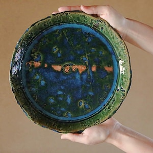 May include: A handmade ceramic plate with a unique, organic shape. The plate features a vibrant color palette of deep blues, greens, and touches of orange. The rim has a textured, uneven edge, adding to its artisanal appeal. A decorative piece for the home.
