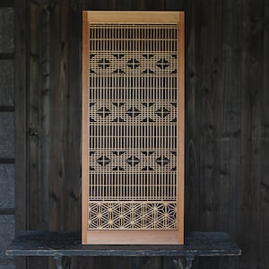 Puede incluir: Un biombo de madera rectangular con un marco de color claro y un intrincado diseño de celosía. La sección superior muestra un patrón geométrico repetitivo de cuadrados y cruces, mientras que la parte inferior presenta un motivo de estrella hexagonal.