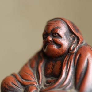 May include: A detailed close-up of a reddish-orange statue of a seated figure with a serene expression. The statue has intricate details, with visible folds in the clothing and a weathered appearance. The figure's face is expressive, with a gentle smile.