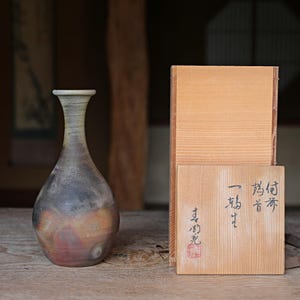 May include: A handcrafted ceramic vase with a slender neck and bulbous body, featuring a blend of gray, brown, and orange hues. Beside it, a wooden box with Japanese calligraphy and a red seal. The items rest on a wooden surface.
