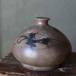 May include: A brown ceramic vase with a rounded body and a narrow neck. The vase features a black painted bird design. The vase is sitting on a wooden surface. The vase is a decorative item.