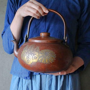 May include: A vintage copper teapot with a handle and lid. The teapot features gold-colored floral designs on its surface. The handle and spout are also copper-colored. The teapot is held by a person wearing a blue shirt and skirt.