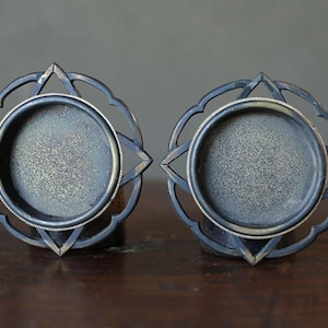 May include: A pair of decorative metal candle holders with a dark, aged finish. Each holder has a circular base within an ornate, openwork design with a star-like shape. The holders are displayed on a dark wooden surface.