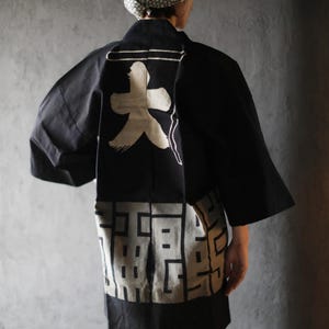 May include: Black kimono-style jacket with white graphic designs on the back. The jacket has wide sleeves and a contrasting geometric pattern at the bottom. A patterned head covering is also visible.