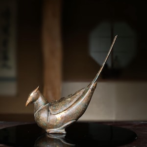May include: A bronze-toned metal bird figurine with a pointed crest, a curved body, and a long, slender tail extending upwards. The surface of the sculpture features a weathered, textured patina with visible dark and metallic brown colour variations.
