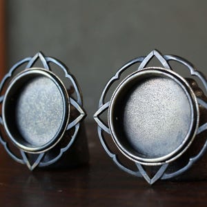 Könnte beinhalten: Zwei silberfarbene Metallkerzenhalter. Jeder hat eine kreisförmige Basis mit einem dekorativen, durchbrochenen Design mit einem Blumen- oder Sternmuster. Die Halter werden auf einer dunklen Holzoberfläche präsentiert.