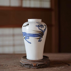 Könnte beinhalten: Eine weiße Keramikvase mit einem handgemalten blauen und goldenen Drachenmotiv, das sich um den Körper windet. Die Vase hat einen schmalen Hals und eine breitere, abgerundete Schulter, die sich zur Basis hin verjüngt und auf einem Holzsockel steht.