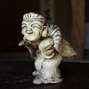 Op de afbeelding: Een vintage, gebroken witte beeldje van een lachende, oudere man in traditionele kledij. De figuur houdt een rond voorwerp vast en heeft zwart-bruine gestreepte accenten. Het beeld staat op een donkere ondergrond.