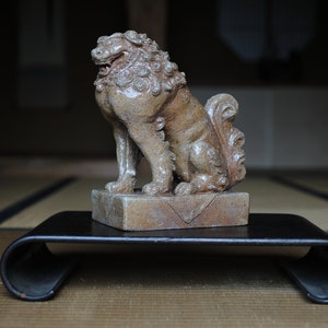 May include: A ceramic lion statue with intricate details, sitting on a black wooden stand. The lion is in a seated position with its head turned to the side.