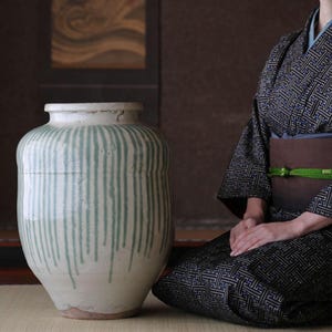 Puede incluir: Una gran jarra de cerámica blanquecina con líneas verticales de esmalte goteado verde claro. La jarra tiene un cuerpo ancho y redondeado y un borde ligeramente ensanchado. El fondo presenta a una persona con un kimono estampado.