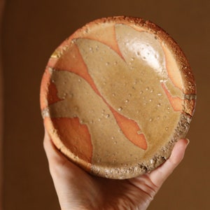 Könnte beinhalten: Handgefertigte Keramikschale mit einzigartigem Design. Die Schale hat einen beigefarbenen Innenraum mit orangefarbenen abstrakten Formen und einer gesprenkelten Oberfläche. Der Rand und die Außenseite haben eine raue Textur. Durchmesser ca. 15 cm.