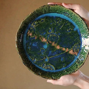 May include: A handmade ceramic plate with a unique design. The plate features a dark green, textured rim and a center with shades of blue and green. The design includes geometric patterns and orange accents. The plate is held by two hands.