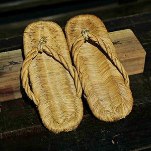 May include: A pair of straw sandals, also known as zori, are made of woven straw and have a simple design. The sandals are light brown in color and have a slightly textured surface.