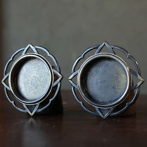 Może przedstawiać: Dwa dekoracyjne metalowe świeczniki. Każdy uchwyt ma okrągłą podstawę ze szczegółową, przypominającą gwiazdę ramą. Metal ma ciemne, postarzane wykończenie, nadające im wygląd vintage. Uchwyty są prezentowane na drewnianej powierzchni.