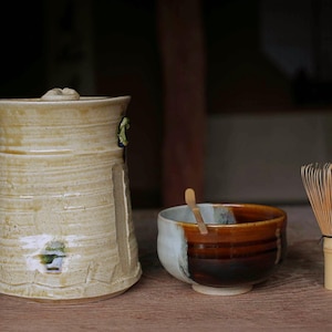 May include: A ceramic tea set featuring a tall, textured beige jar with a lid, a two-toned tea bowl with a wooden spoon, and a bamboo whisk. The set is arranged on a wooden surface.