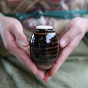May include: A small, barrel-shaped ceramic jar with a glossy, dark brown and black mottled glaze. The jar features a smooth, light-colored circular lid at the top and a matte, unglazed tan base, showcasing a distinct contrast in texture and color.