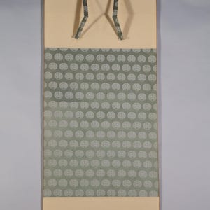Può includere: Un rotolo di pittura giapponese tradizionale con uno sfondo color crema e una sezione a motivi al centro. Il motivo presenta ripetuti disegni circolari bianchi su uno sfondo verde tenue. Il rotolo ha un meccanismo di sospensione nella parte superiore.
