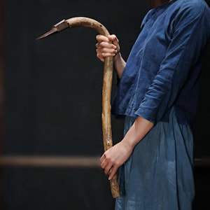 Könnte beinhalten: Eine Person hält ein langes, gebogenes Holzwerkzeug mit einer Metallspitze. Das Werkzeug ist braun und hat eine natürliche Oberfläche. Die Person trägt ein blaues Hemd und eine blaue Hose.