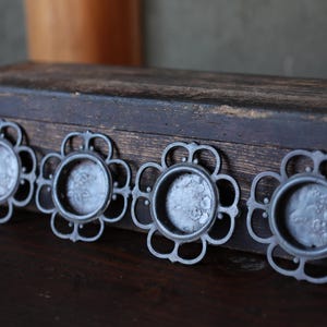 Puede incluir: Cuatro tiradores de cajón antiguos de metal plateado con diseño floral. Cada tirador tiene un centro circular y un borde floral calado decorativo. Los tiradores están dispuestos en fila sobre una superficie de madera oscura.