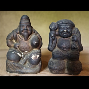 May include: Two antique, seated Asian figurines. The figurine on the left is a dark brown colour with a long beard and a hat. The figurine on the right is black and holds a staff and a bag. Both have smiling faces and are weathered.