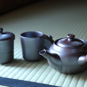Puede incluir: Una tetera de cerámica marrón con una superficie texturizada y una tapa a juego, una taza de cerámica marrón y un recipiente de cerámica marrón con tapa. Los artículos están sobre una estera verde claro.