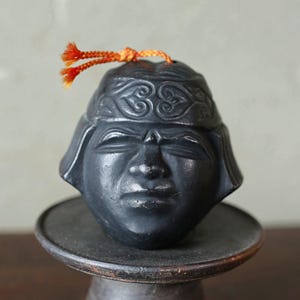 May include: A dark gray, stylized head sculpture with closed eyes, atop a round, dark gray pedestal. The head has a decorative helmet and a small orange and yellow tassel. The sculpture is likely a decorative item.