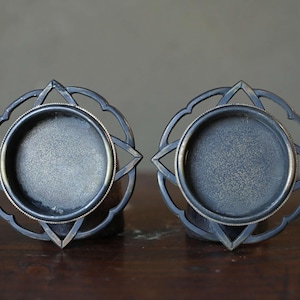 May include: Two decorative metal candle holders. Each holder features a circular base surrounded by an ornate, openwork design. The metal has a dark, aged appearance, with a silver-colored rim around the candle cup. The holders are displayed on a wooden surface.