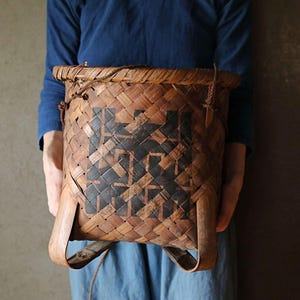 May include: A woven brown basket with black geometric designs. The basket has two leather straps and is being held by a person.